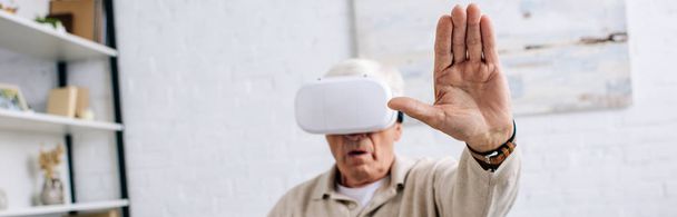 панорамный снимок пожилого человека в гарнитуре виртуальной реальности с протянутой рукой в квартире
  - Фото, изображение