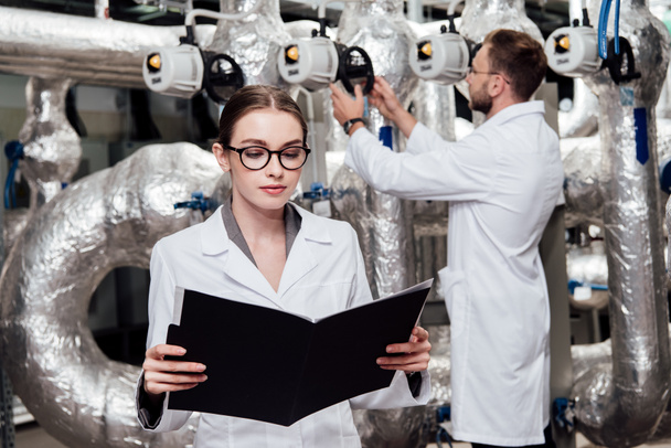 selective focus of attractive engineer in white coat looking at folder near coworker and compressed air system  - Photo, Image