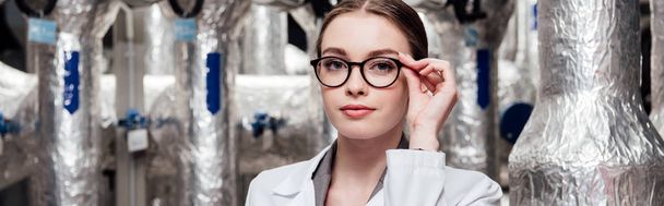 panoramic shot of attractive engineer in white coat touching glasses near air compressed system  - 写真・画像