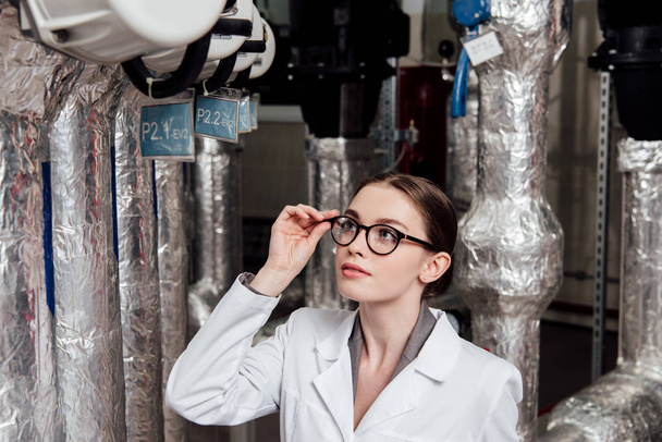 attractive engineer in white coat touching glasses while looking at air compressed system  - 写真・画像