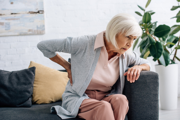 vanhempi nainen, jolla on selkäkipu ja istuu sohvalla asunnossa
  - Valokuva, kuva