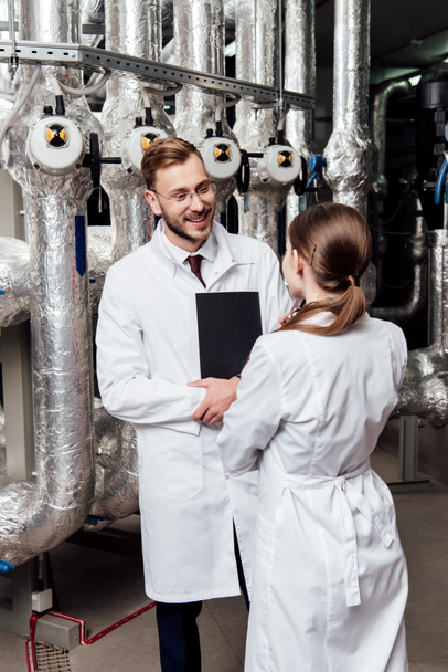 cheerful engineer looking at coworker while standing near air compressor system  - Photo, Image