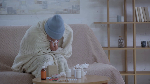 παχύς άνθρωπος κάθεται κοντά στο τραπέζι με φάρμακα, φτάρνισμα και περιτύλιγμα σε κουβέρτα  - Πλάνα, βίντεο