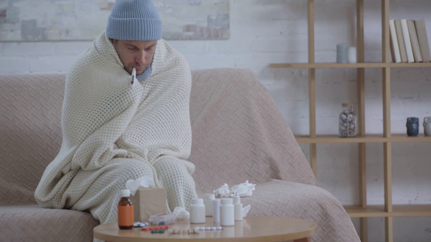 chory człowiek siedzący przy stole z lekami i pomiarem temperatury - Materiał filmowy, wideo