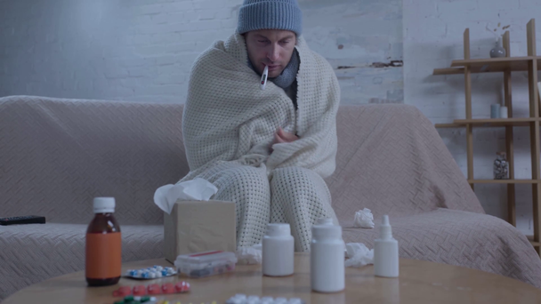 άρρωστος άνθρωπος κάθεται κοντά στο τραπέζι με τα φάρμακα και τη μέτρηση της θερμοκρασίας - Πλάνα, βίντεο