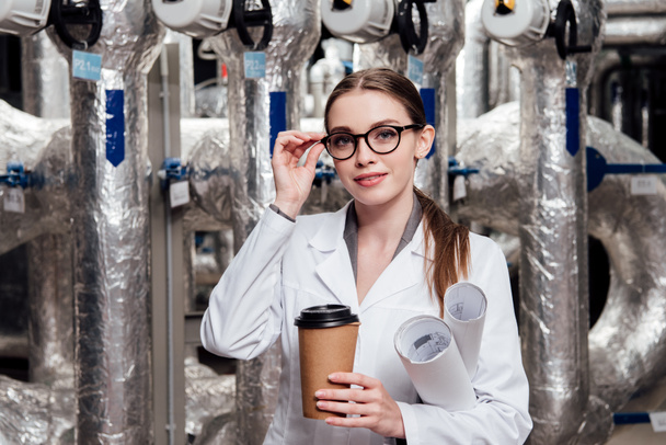 cheerful engineer in white coat and glasses holding blueprints and paper cup near air supply system  - Photo, Image