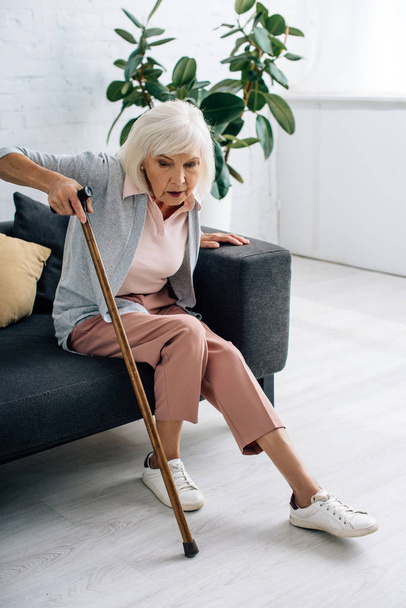 senior woman with wooden cane sitting on sofa in apartment  - 写真・画像