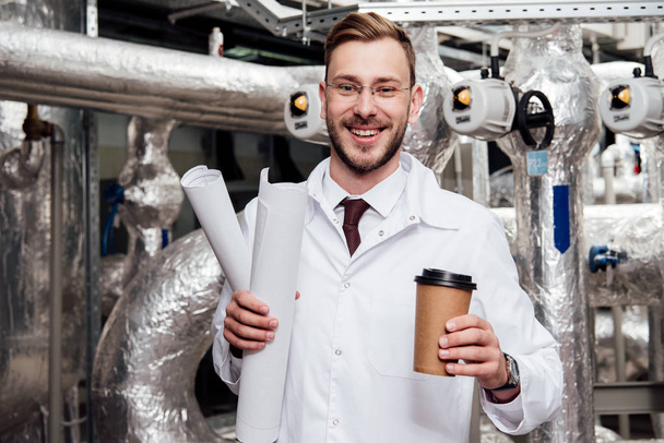 веселый инженер в белом халате держит чертежи и бумажные чашки возле системы подачи воздуха
  - Фото, изображение