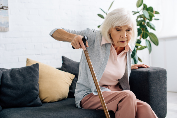 senior woman with wooden cane sitting on sofa in apartment  - Foto, imagen