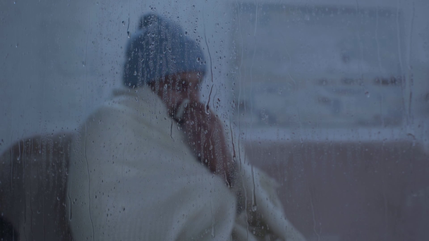 sick man sitting near window glass with raindrops and sneezing into napkin - 映像、動画