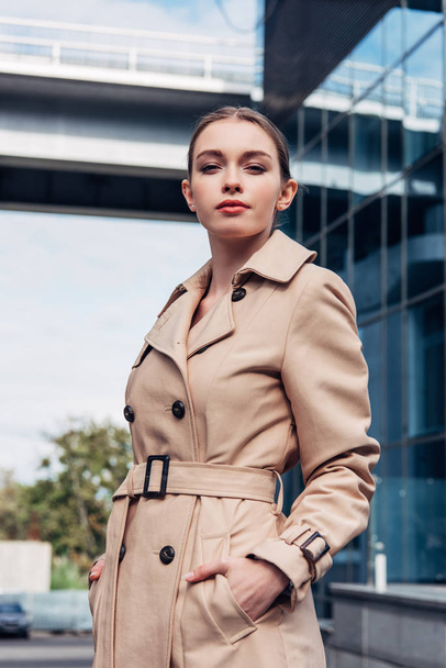 atraktivní žena v trenč kabát stojící s rukama v kapsách  - Fotografie, Obrázek