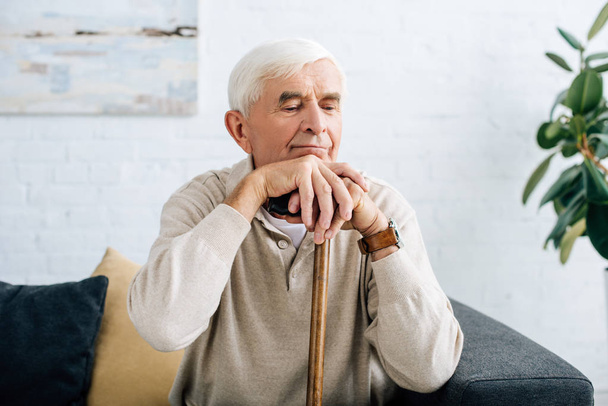senior man sitting on sofa and holding wooden cane in apartment  - Foto, afbeelding
