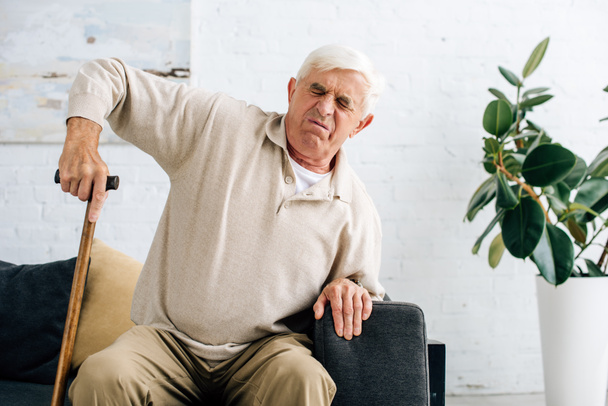 ηλικιωμένος άνδρας που έχει πόνο και κρατώντας ξύλινο μπαστούνι στο διαμέρισμα  - Φωτογραφία, εικόνα