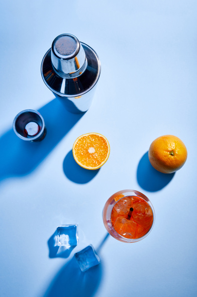 vista superior do coquetel Aperol Spritz, laranjas, coqueteleira, cubos de gelo e copo de medição no fundo azul
  - Foto, Imagem