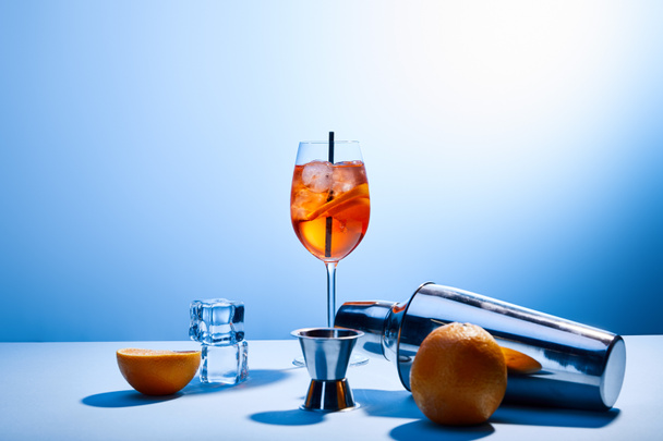 κοκτέιλ Aperol Spritz, πορτοκάλια, σέικερ, παγάκια και κύπελλο μέτρησης σε μπλε φόντο  - Φωτογραφία, εικόνα