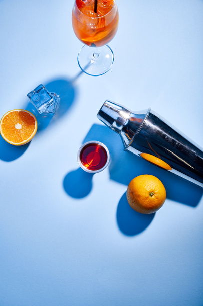 vrchní pohled na Aperol Spritz, pomeranče, třepačka, kostky ledu a odměrka na modrém pozadí  - Fotografie, Obrázek