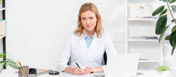 πανοραμική λήψη του ελκυστικού διατροφολόγου κάθεται στο τραπέζι και κρατώντας στυλό στην κλινική  - Φωτογραφία, εικόνα
