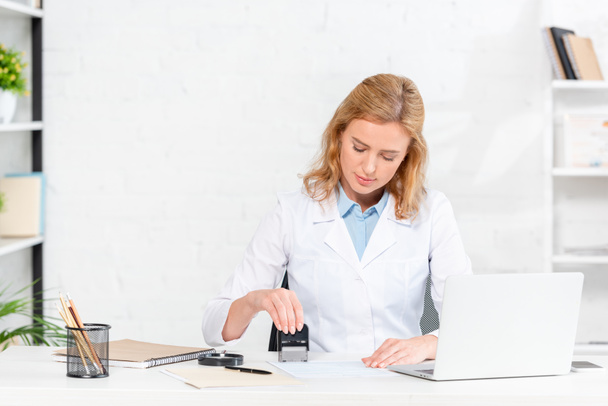 attractive nutritionist sitting at table and stamping paper in clinic  - Foto, Bild