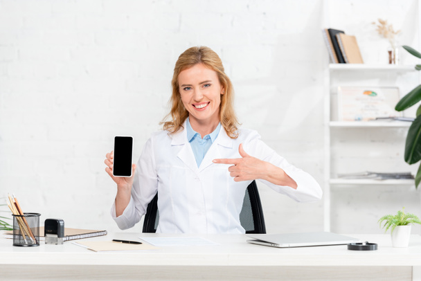 uśmiechnięty dietetyk siedzący przy stole i wskazujący palcem na smartfona z przestrzenią do kopiowania w klinice  - Zdjęcie, obraz