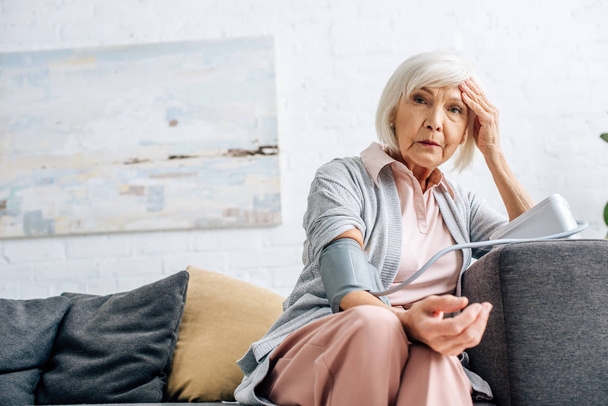 Seniorin sitzt auf Sofa und misst Blutdruck in Wohnung  - Foto, Bild