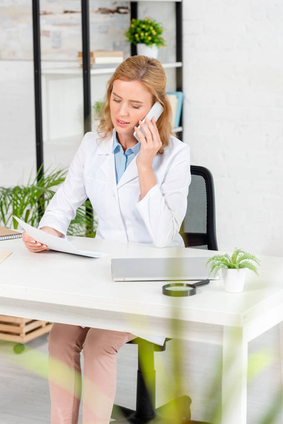 attractive nutritionist holding paper and talking on smartphone in clinic  - Foto, imagen