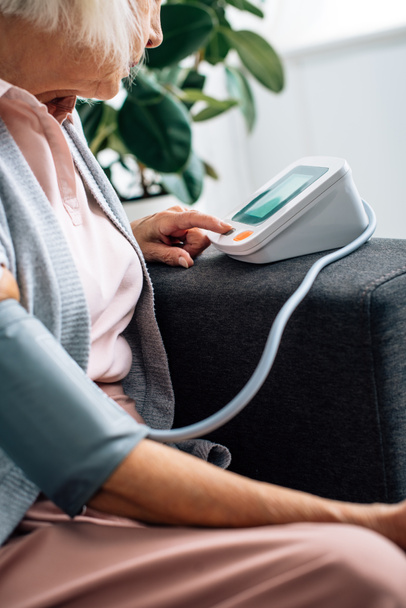 bijgesneden zicht op oudere vrouw zittend op de bank en het meten van de bloeddruk in appartement  - Foto, afbeelding