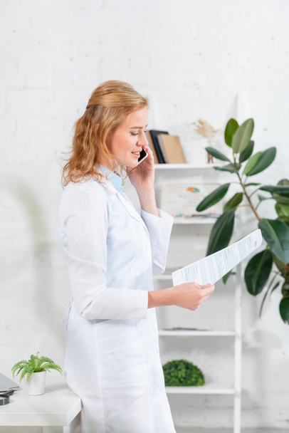 side view of attractive nutritionist holding paper and talking on smartphone in clinic  - Фото, зображення