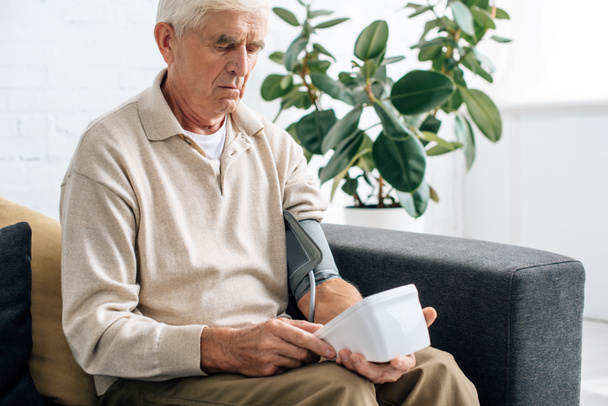 starszy mężczyzna mierzy ciśnienie krwi i siedzi na kanapie w mieszkaniu  - Zdjęcie, obraz