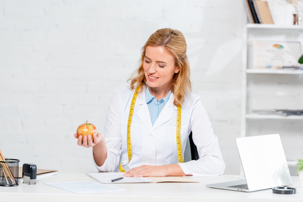 nutritionniste souriant assis à la table et tenant pomme à la clinique
  - Photo, image