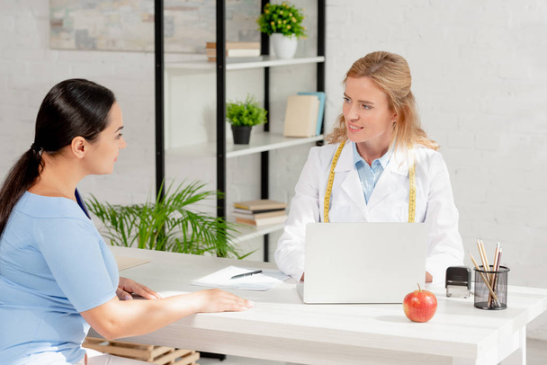 smiling nutritionist sitting at table and talking with patient in clinic  - Photo, Image