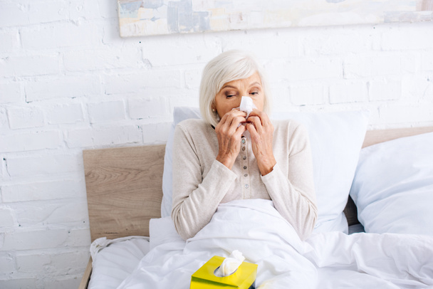 ill senior woman sneezing and holding napkin in bed  - Photo, Image