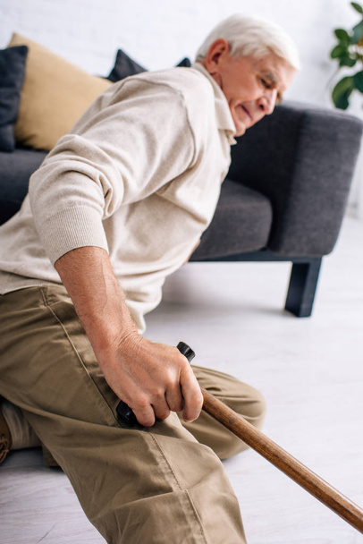 selective focus of senior man holding wooden cane and feeling pain in apartment  - Fotoğraf, Görsel