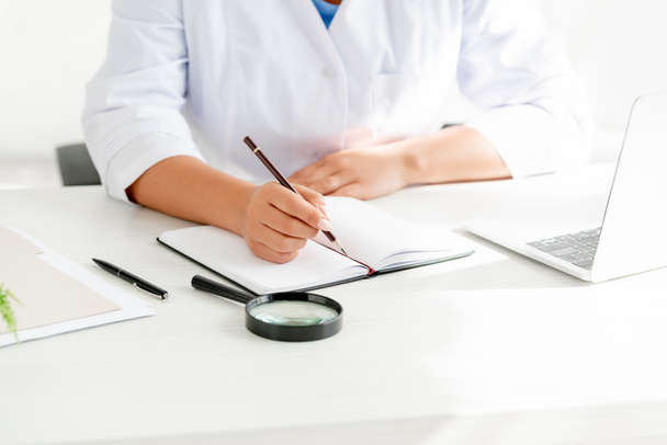 vista recortada del dermatólogo sentado en la mesa y la escritura en el cuaderno en la clínica
  - Foto, Imagen