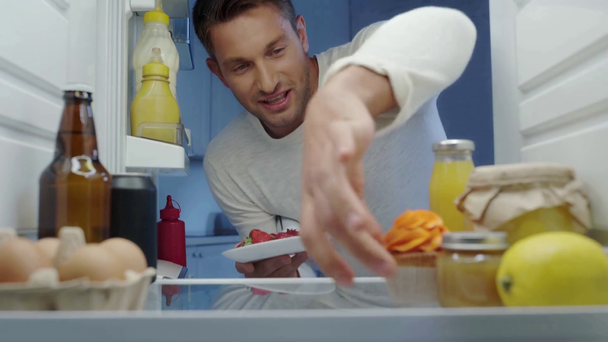 hongerige man neemt aardbeien en container van slagroom uit de koelkast - Video