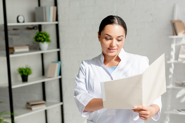 attractive and smiling dermatologist in white coat holding folder in clinic  - Foto, Imagen