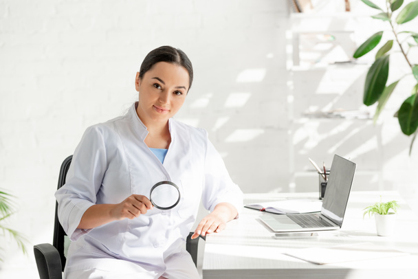 attractive dermatologist sitting at table and holding magnifying glass in clinic  - Photo, Image