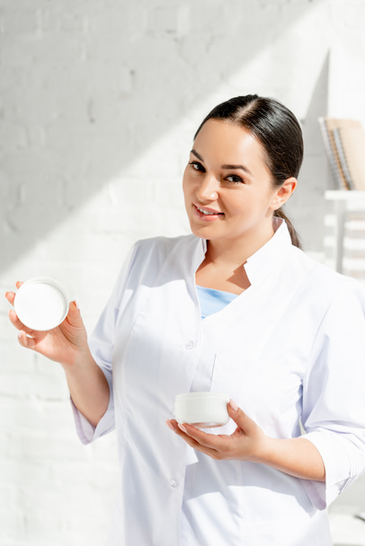 dermatólogo sonriente sosteniendo crema cosmética y mirando a la cámara en la clínica
  - Foto, Imagen