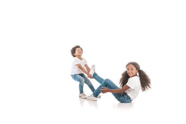 χαρούμενο αφροαμερικανό αγόρι σέρνει την αδελφή του από το πόδι σε λευκό φόντο - Φωτογραφία, εικόνα