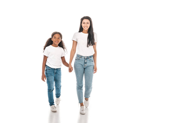 feliz afroamericano madre e hija cogidas de la mano y mirando a la cámara mientras camina sobre fondo blanco
 - Foto, Imagen