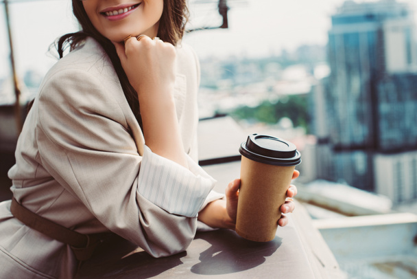 cropped view of smiling girl in beige suit posing on roof with coffee to go - Photo, image