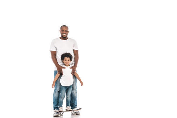happy african american man supporting cheerful son standing on penny board on white background - Фото, изображение