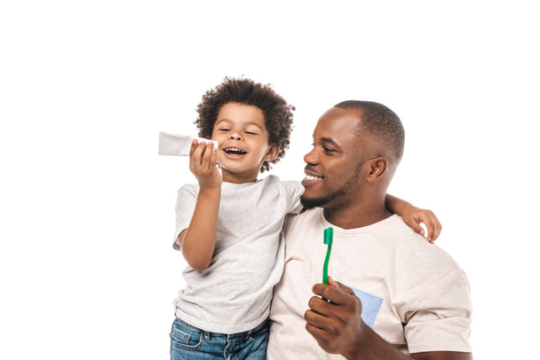 alegre afroamericano chico mirando dentífrico cerca feliz padre sosteniendo cepillo de dientes aislado en blanco
 - Foto, Imagen