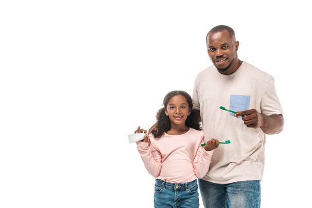 χαρούμενο αφροαμερικάνικο παιδί που κρατάει οδοντόβουρτσα και οδοντόκρεμα ενώ στέκεται δίπλα στον πατέρα κρατώντας οδοντόβουρτσα που είναι απομονωμένη στα λευκά - Φωτογραφία, εικόνα