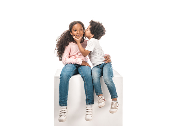 cute african american boy kissing adorable sister while sitting on white cube together isolated on white - Fotoğraf, Görsel