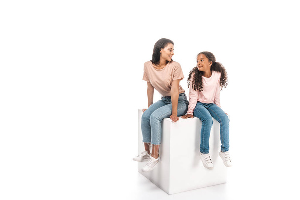 smiling african american mother and daughter looking at each other while sitting on white cube on white background - Φωτογραφία, εικόνα