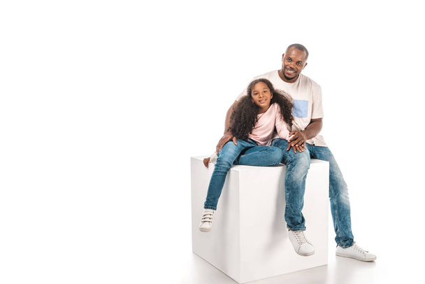 smiling african american father and daughter looking at camera while sitting on cube together on white background - Photo, image