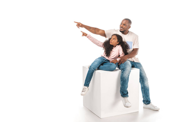 sorrindo afro-americano pai e filha olhando para longe e apontando com o dedo enquanto sentados no cubo juntos no fundo branco
 - Foto, Imagem