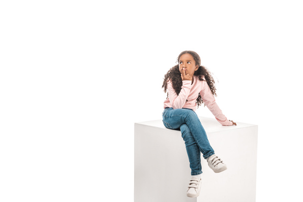 thoughtful african american child looking away while sitting on white cube isolated on white - Foto, Imagem
