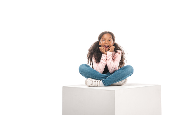 cheerful african american child sitting on white cube with crossed legs isolated on white - Zdjęcie, obraz