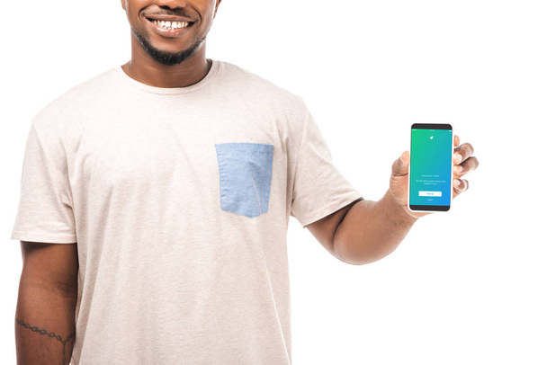 KYIV, UCRANIA - 15 de agosto de 2019: Vista parcial del hombre afroamericano sonriente sosteniendo el teléfono inteligente con la aplicación de Twitter en la pantalla aislada en blanco
. - Foto, Imagen